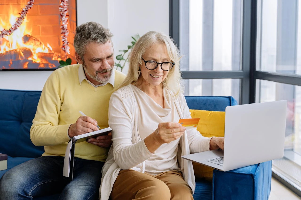 A couple in their 40s smiling while reviewing retirement plans on a laptop, with a scenic beach visible through a window.